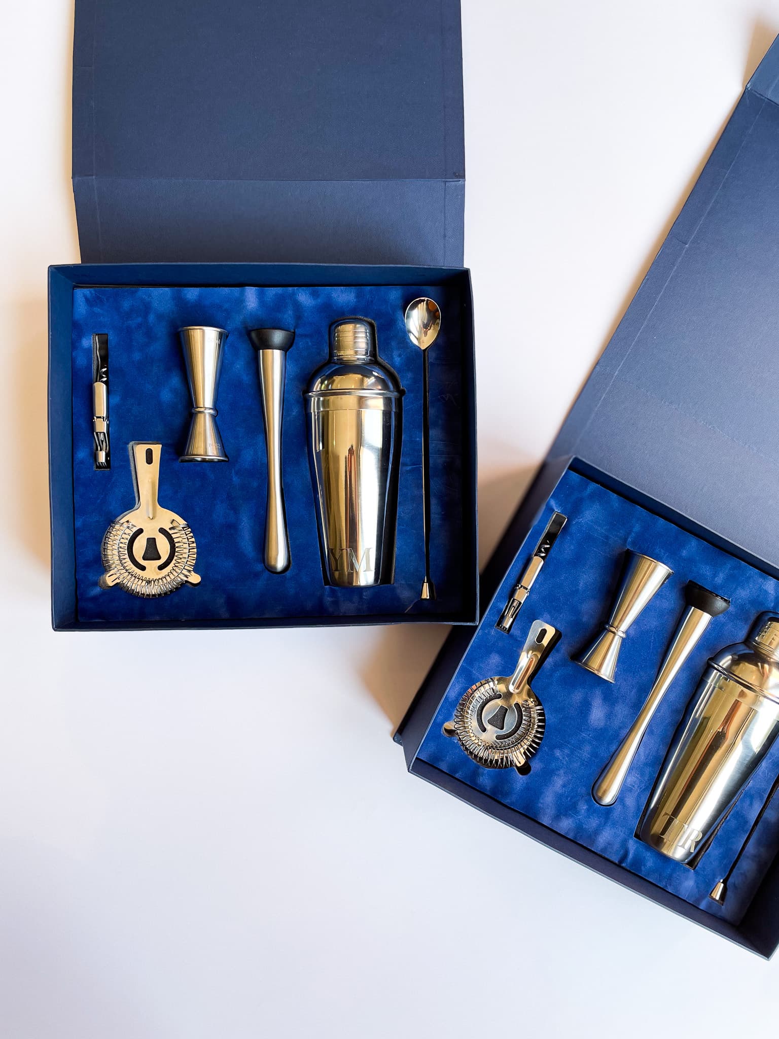 Two open navy blue gift boxes with deep blue velvet lining containing gold cocktail shaker, jigger, muddler, bar strainer and mixing spoon