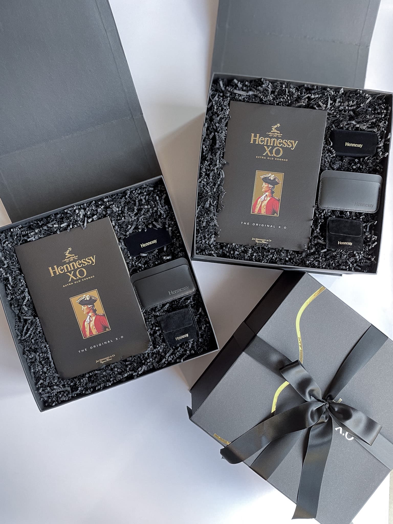 Two open dark grey gift boxes containing Hennessy XO presentation books and branded merchandise, with a ribbon-wrapped box in the foreground