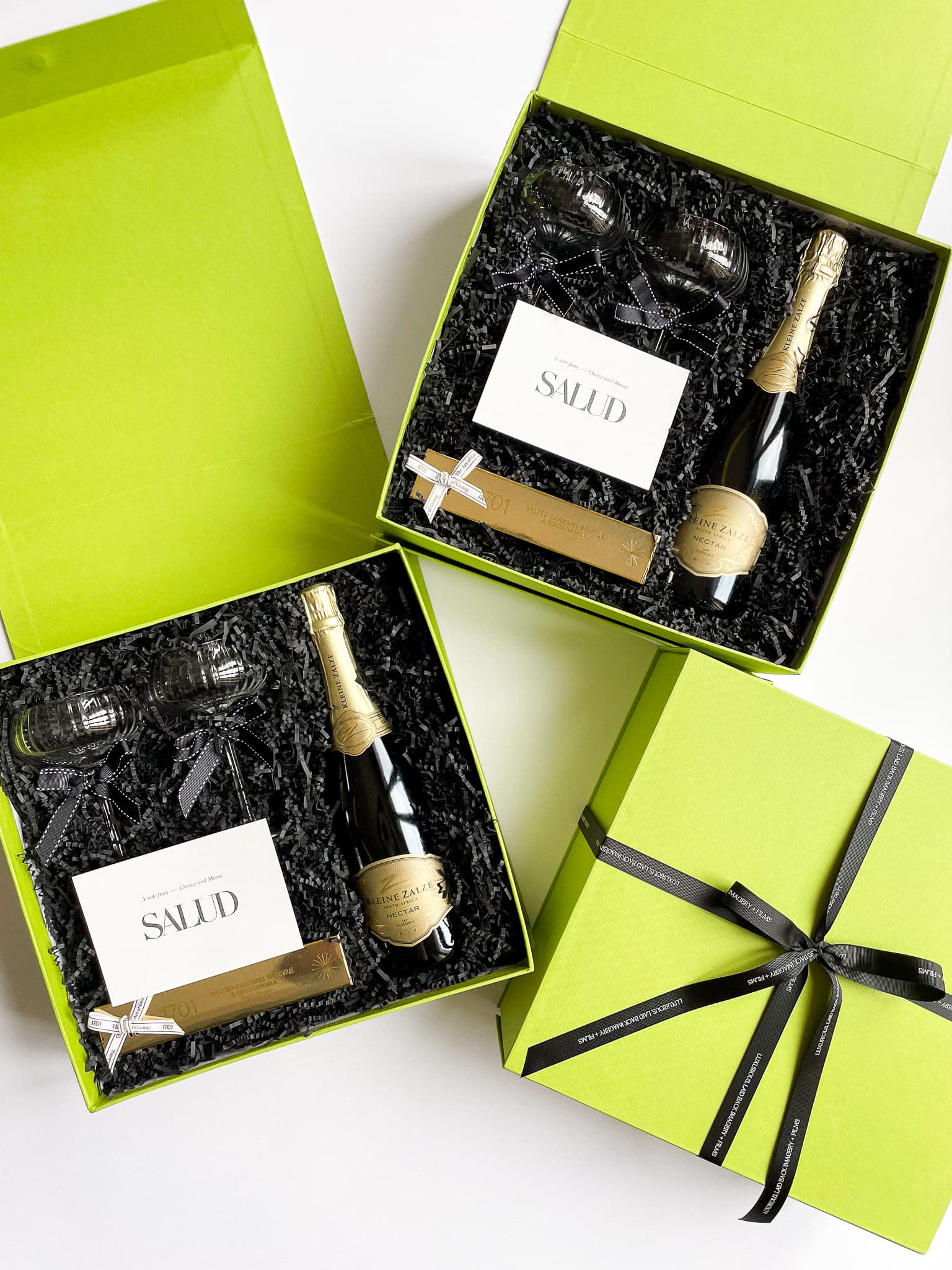 Three lime green gift boxes open to reveal black crinkle paper with Kleine Zalze champagne bottles, champagne glasses and branded Salud cards, with one box tied with a black ribbon