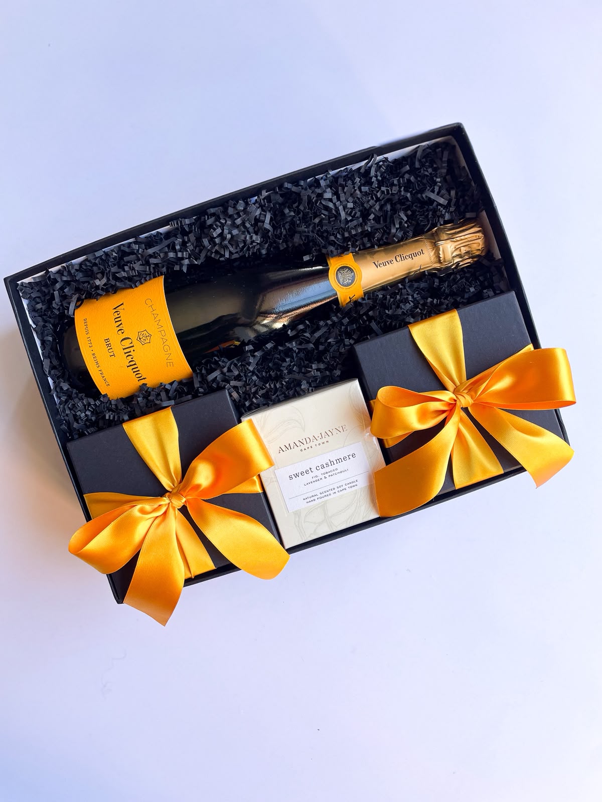 Open black gift box with black crinkle paper containing a Veuve Clicquot champagne bottle, Amanda Jayne sweet cashmere candle and wrapped gifts tied with orange satin ribbons